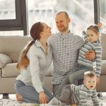 Cheerful mother and father smiling at each other while little cute boy crawling on floor and preschool girl in striped turtleneck raising arm and looking at camera