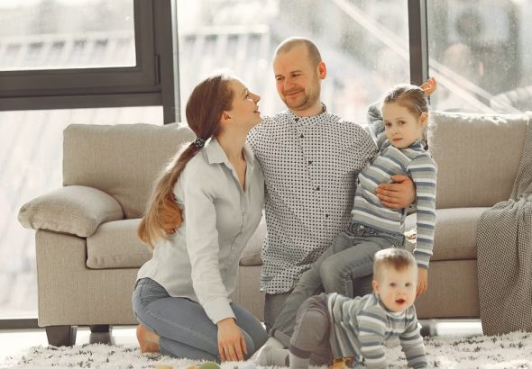 Cheerful mother and father smiling at each other while little cute boy crawling on floor and preschool girl in striped turtleneck raising arm and looking at camera