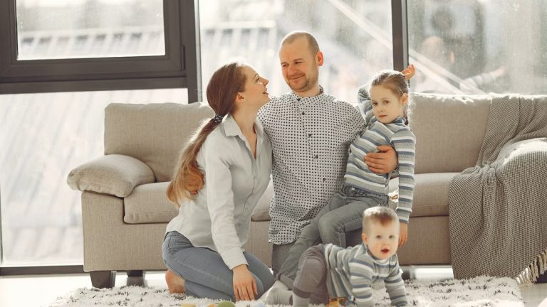 Cheerful mother and father smiling at each other while little cute boy crawling on floor and preschool girl in striped turtleneck raising arm and looking at camera